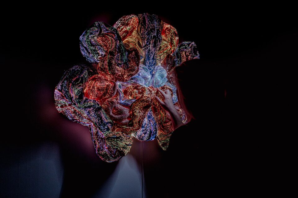 Glowing multicoloured textile sculpture suspended in a dark room at ANDETAG Stockholm