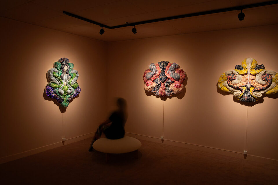 Three glowing textile wall sculptures in a warm-lit gallery at ANDETAG Stockholm with a visitor on a round ottoman