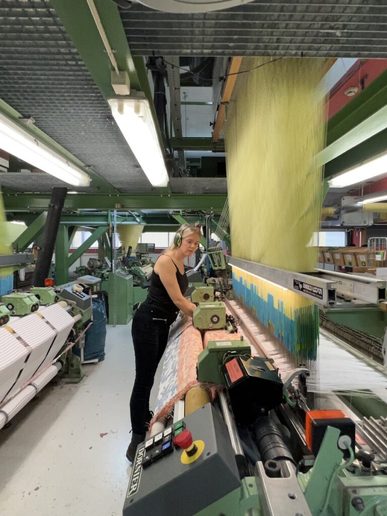 Malin Tadaa at a large-format jacquard loom in the textile factory weaving optical fibre textile with yellow and blue warp threads