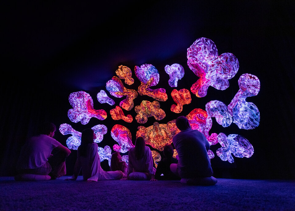 Four visitors seated together before a large wall of luminous textile sculptures at ANDETAG.