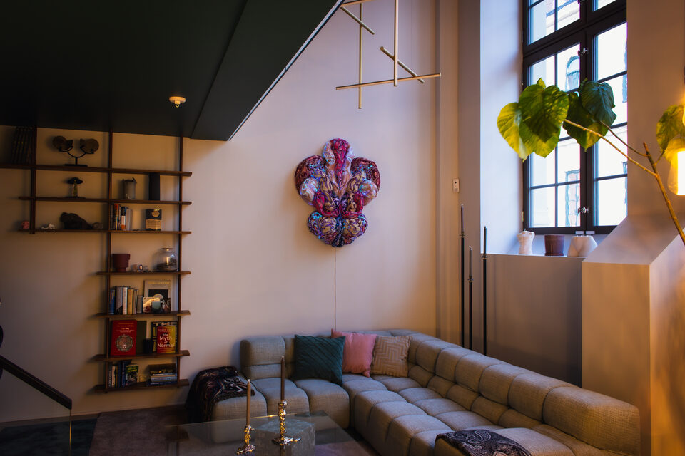 The artwork ANDETAG no. 12 in pink and magenta tones hangs above a beige sofa in a daylit living room, with bookshelves on the left and a green plant by the window.