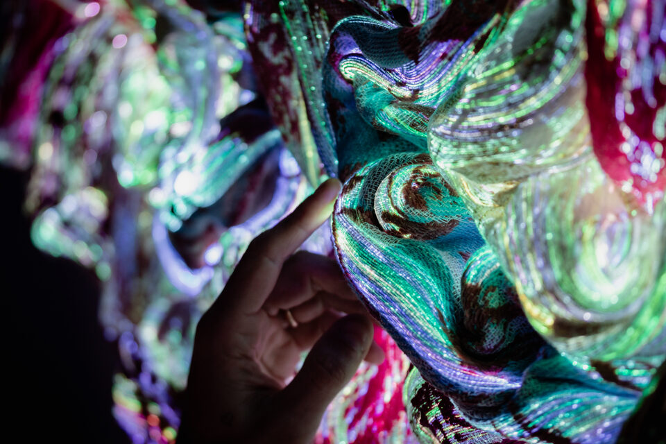 Close-up with a hand pointing toward the artwork Andetag no. 16, green and silver shimmering optical fibre textile.