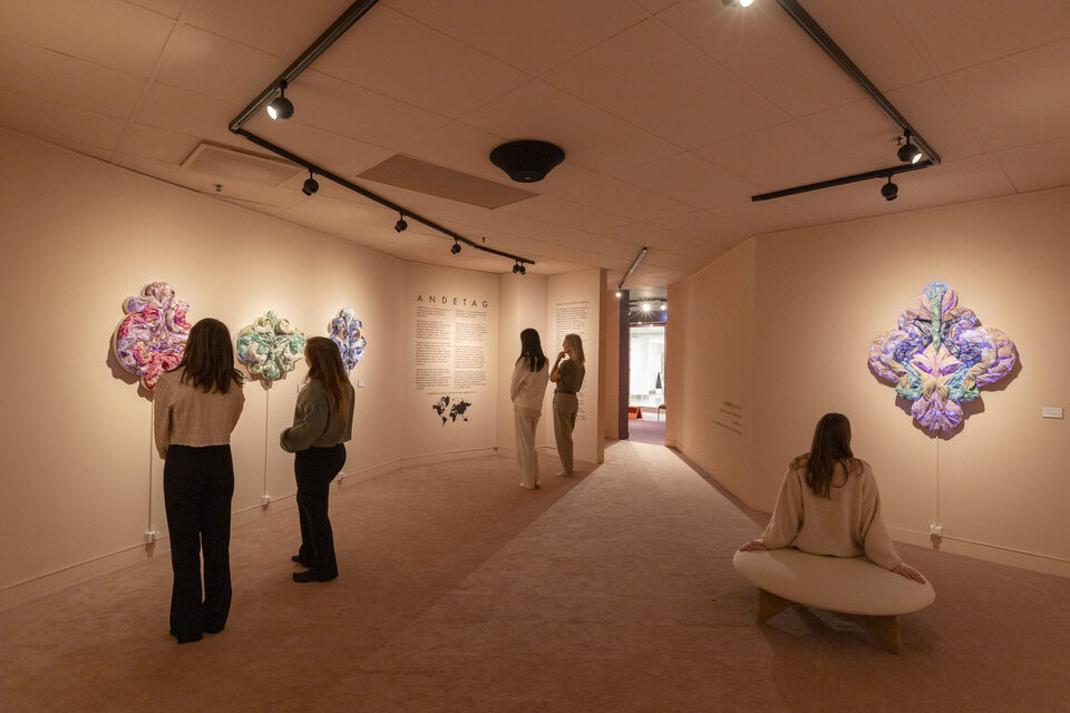 The exhibition gallery with the ANDETAG info panel on the wall and visitors viewing textile works.