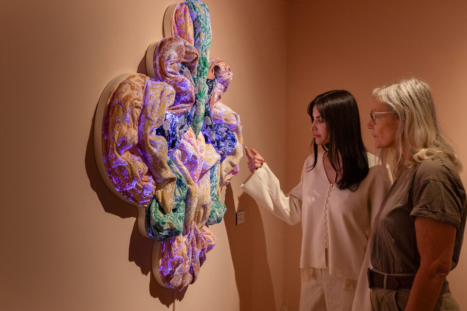 Two visitors contemplating a shimmering textile sculpture in violet, gold, and green.