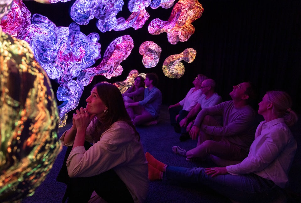 Visitors sitting still with hands clasped near their chins before glowing textile sculptures.