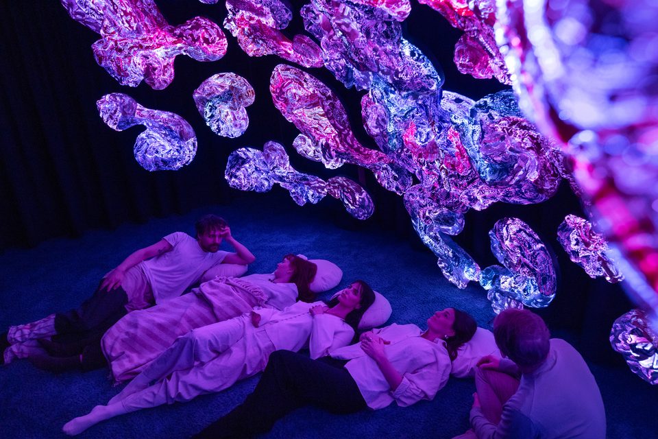 Visitors resting on pillows beneath floating, luminous textile sculptures.