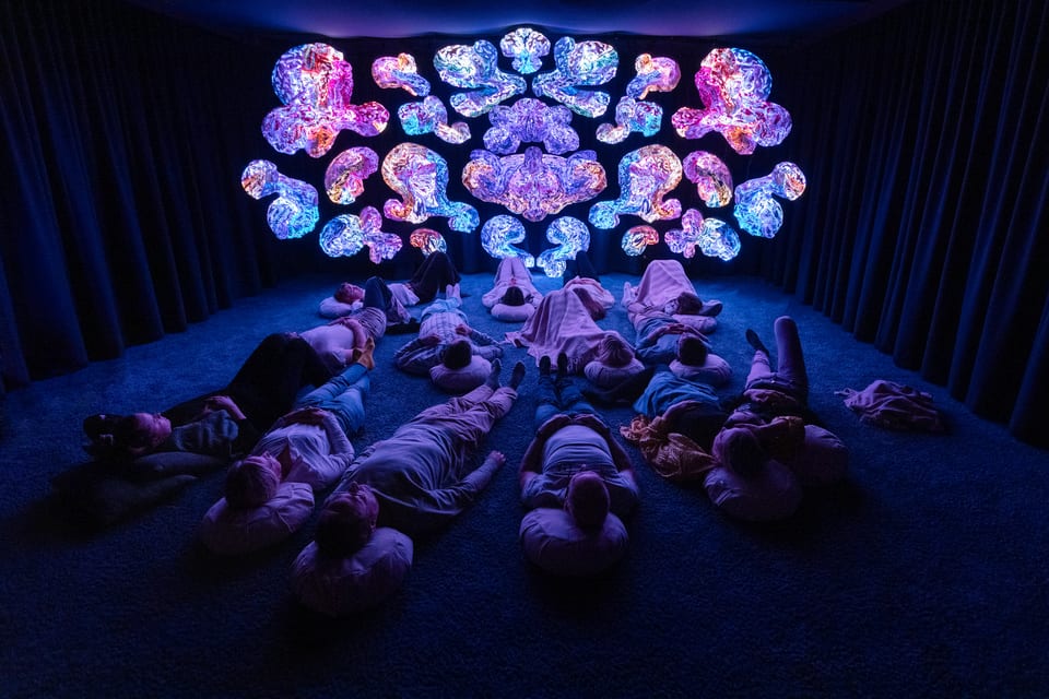 Wide view of the main room at ANDETAG, visitors resting before a wall of breathing light art.