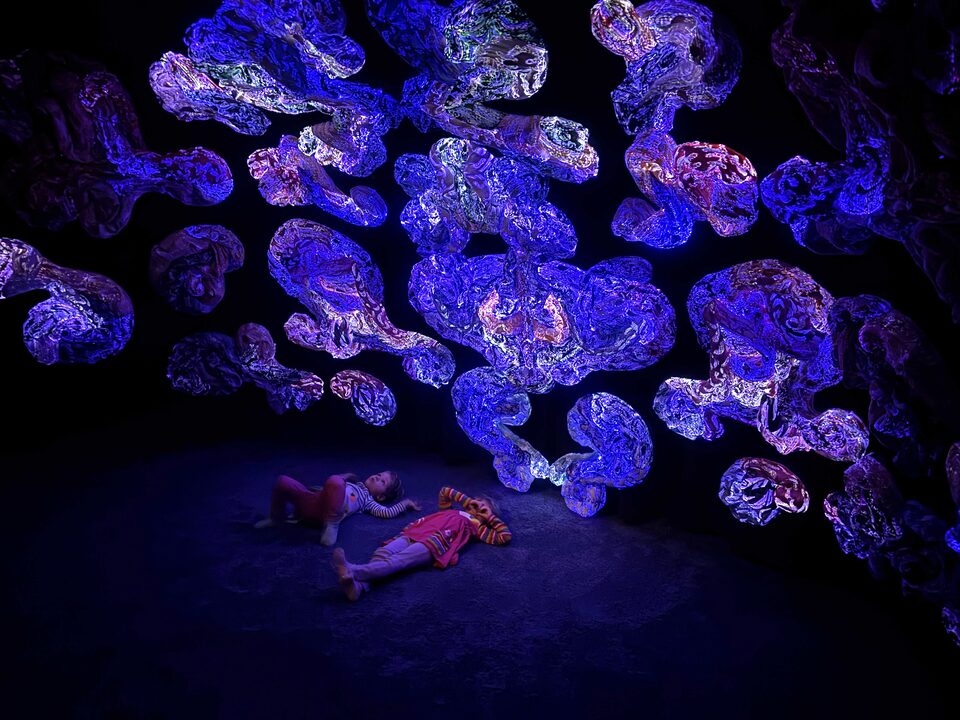 Two children lying on the floor surrounded by large, luminous Andetag sculptures in blue and violet.