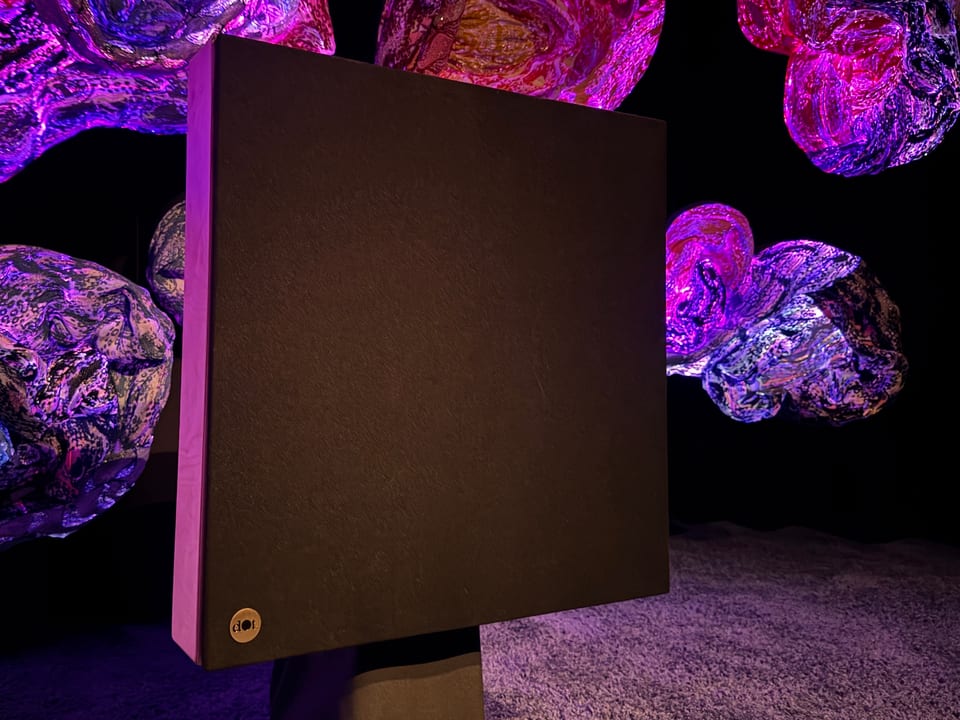 Dark, matte dotAudio speaker panel among luminous textile sculptures in violet and pink light.